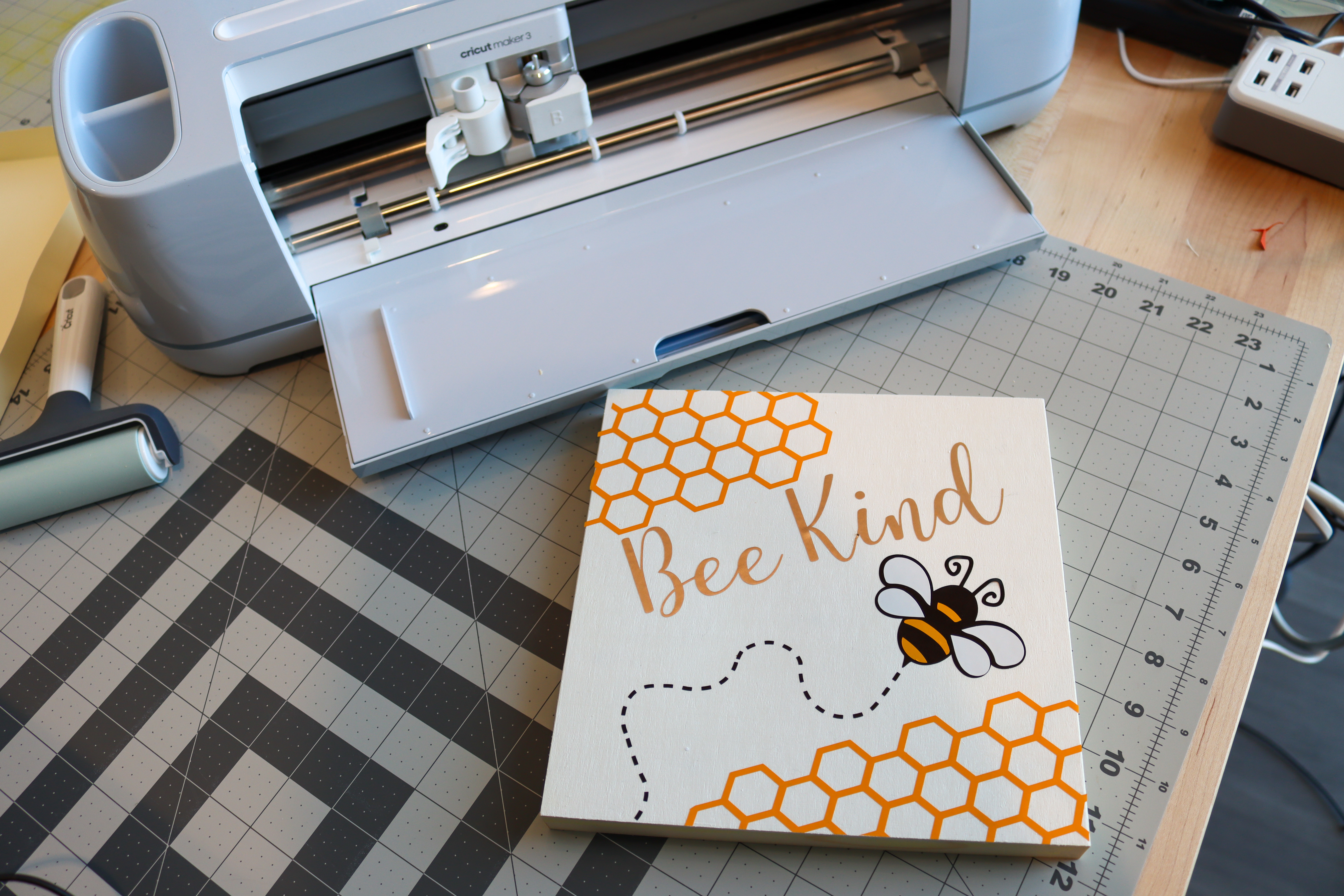Square wooden sign with a honeycomb design on the top left and bottom right corners. A bee is flying across the sign with text that says bee kind in script across the middle of the sign.