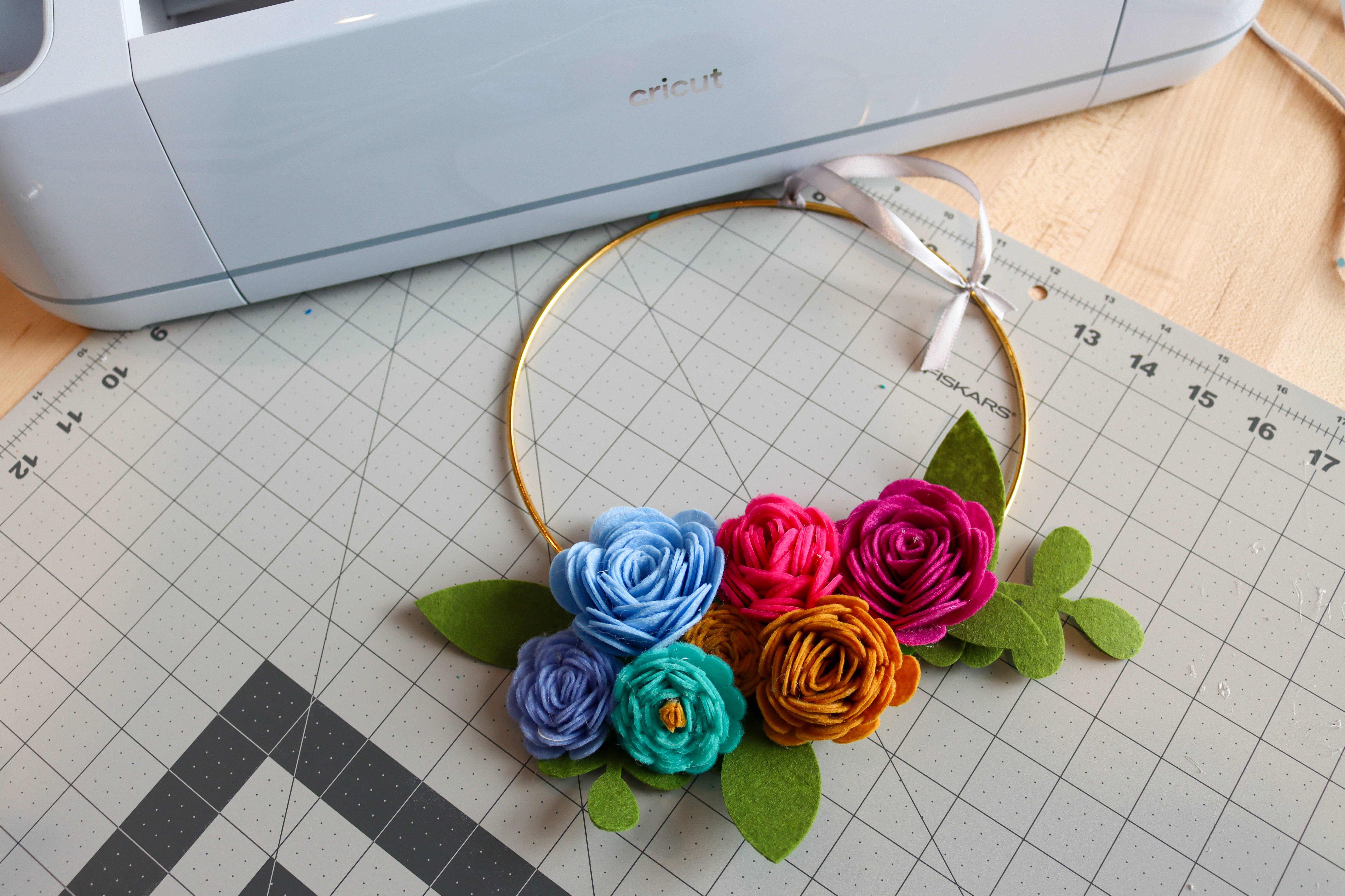 Wreath on a table next to a Circut machine. The wreath is made of a gold hoop with a ribbon tied at the top. It is decorated at the bottom with colorful felt flowers and leaves.