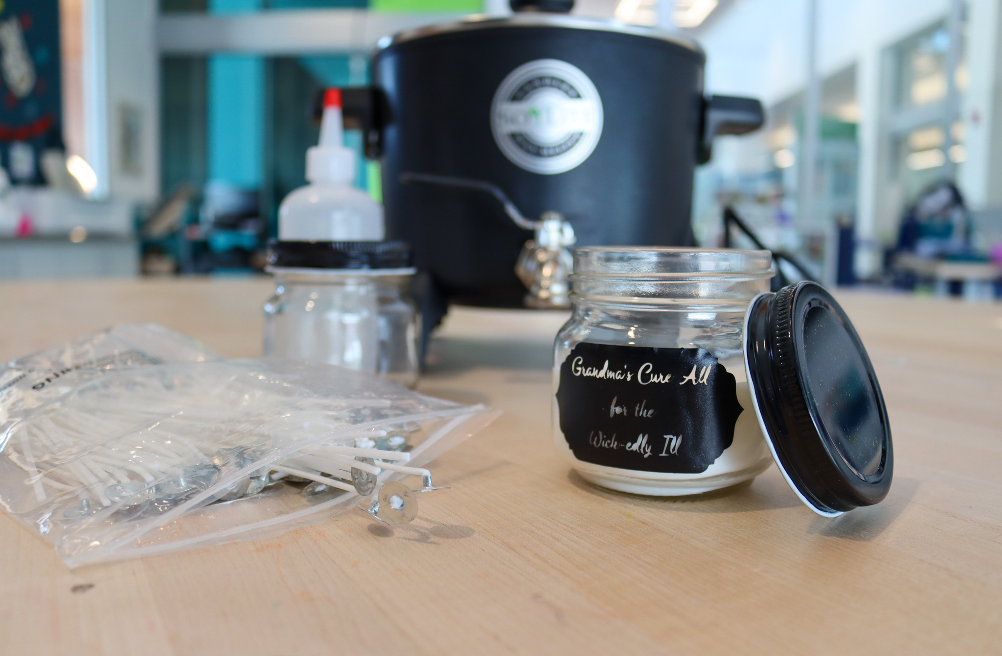 Candle making supplies on a table in Studio 411. A small white wax candle in a glass jar with a black label sits in front of a bag of wicks, a squeeze bottle, and a wax melter.