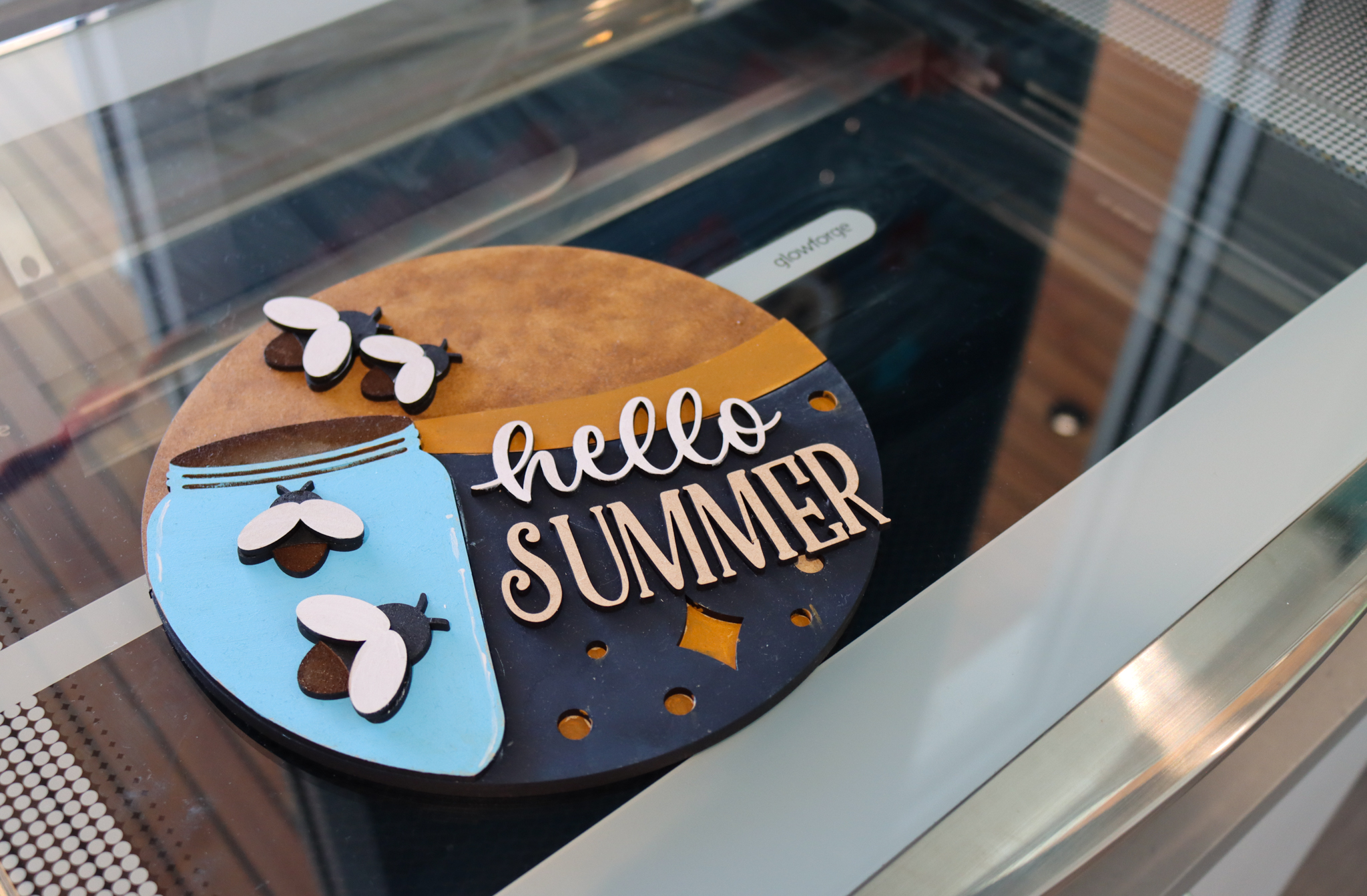 Circular wooden sign that says hello summer in white script next to a blue jar of fireflies