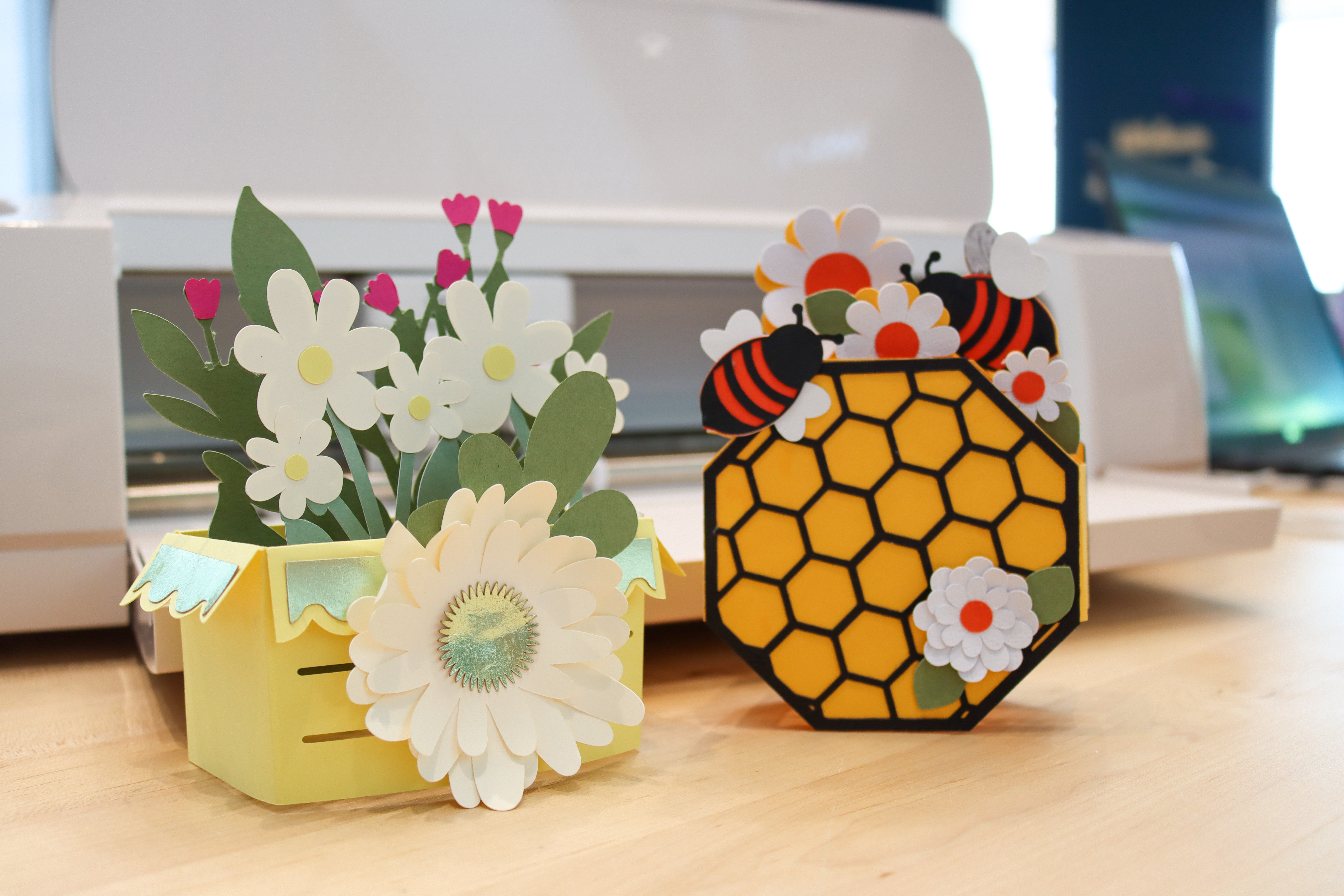 A yellow flower box with white flowers growing from it, all made of paper, next to a decorative beehive with bees and flowers attached, also made of paper.