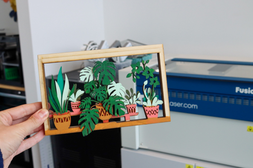 Wood cut in the shapes of potted plants and assembled into a rectangular art piece.