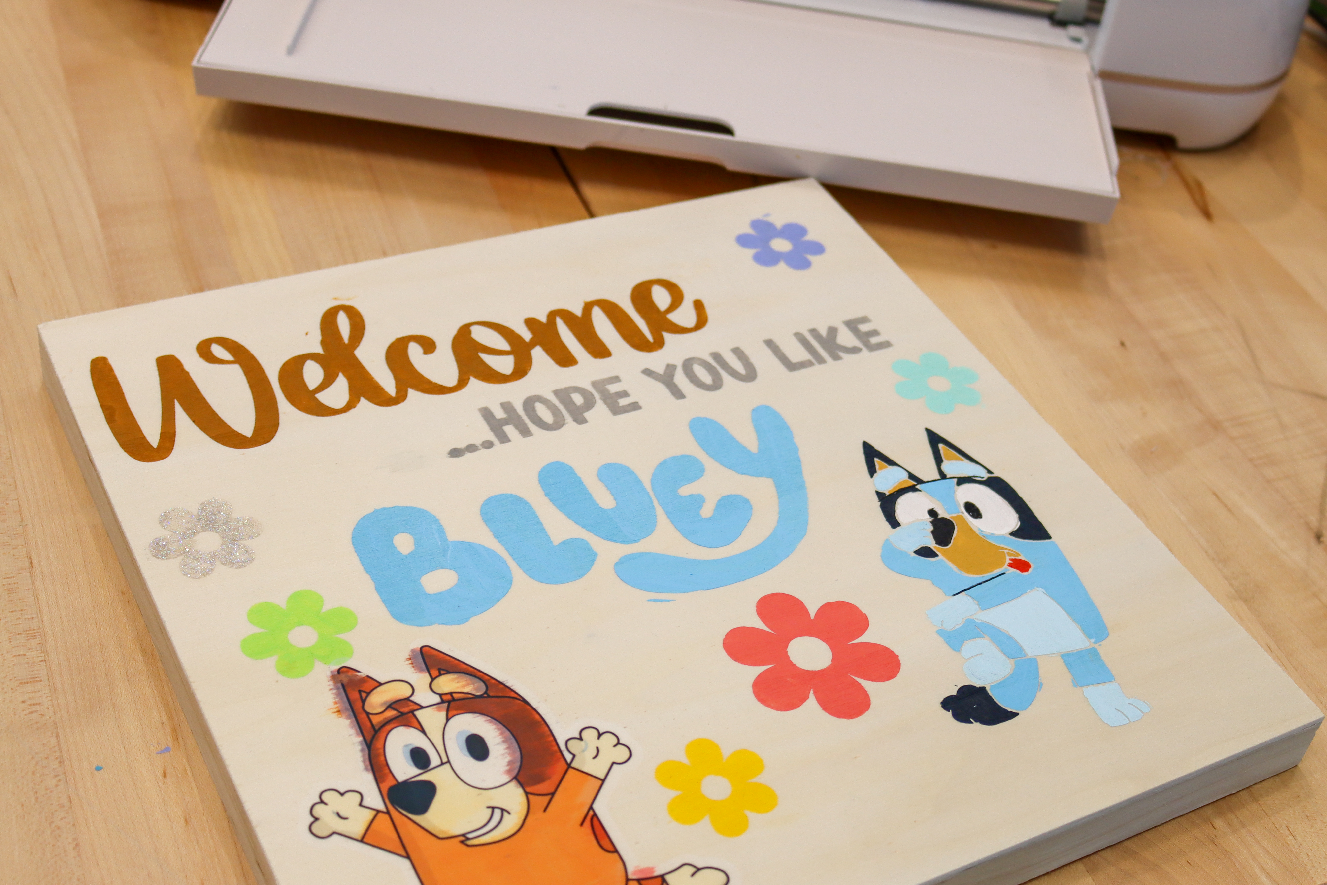Square wooden sign with colorful words painted that say welcome, hope you like Bluey with cartoon characters Bluey and Bingo underneath surrounded by flowers