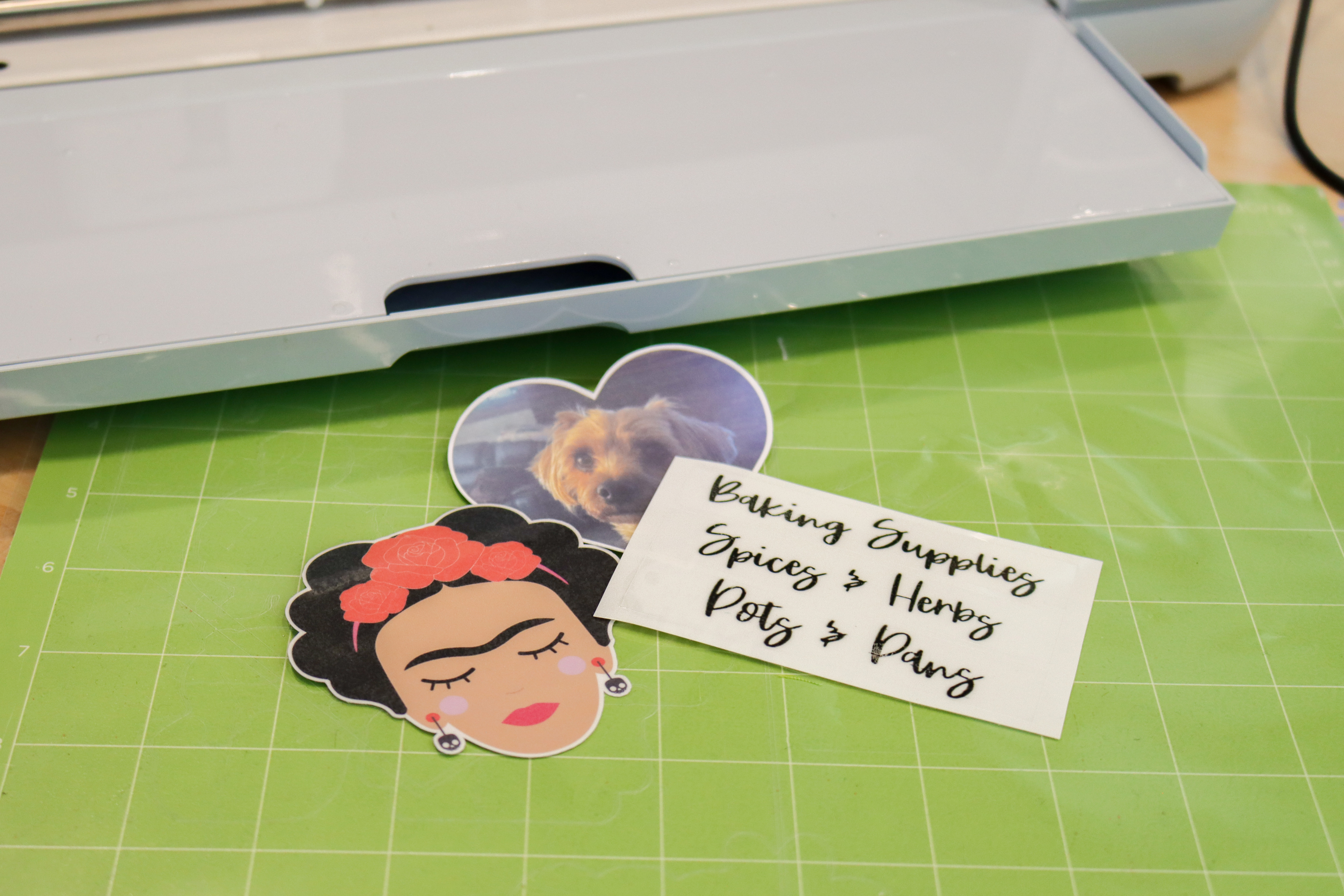 Three stickers on a table in front of a Circut machine. Once sticker is of Frida Kahlo, one is of a dog, and the other are labels for home organizing.