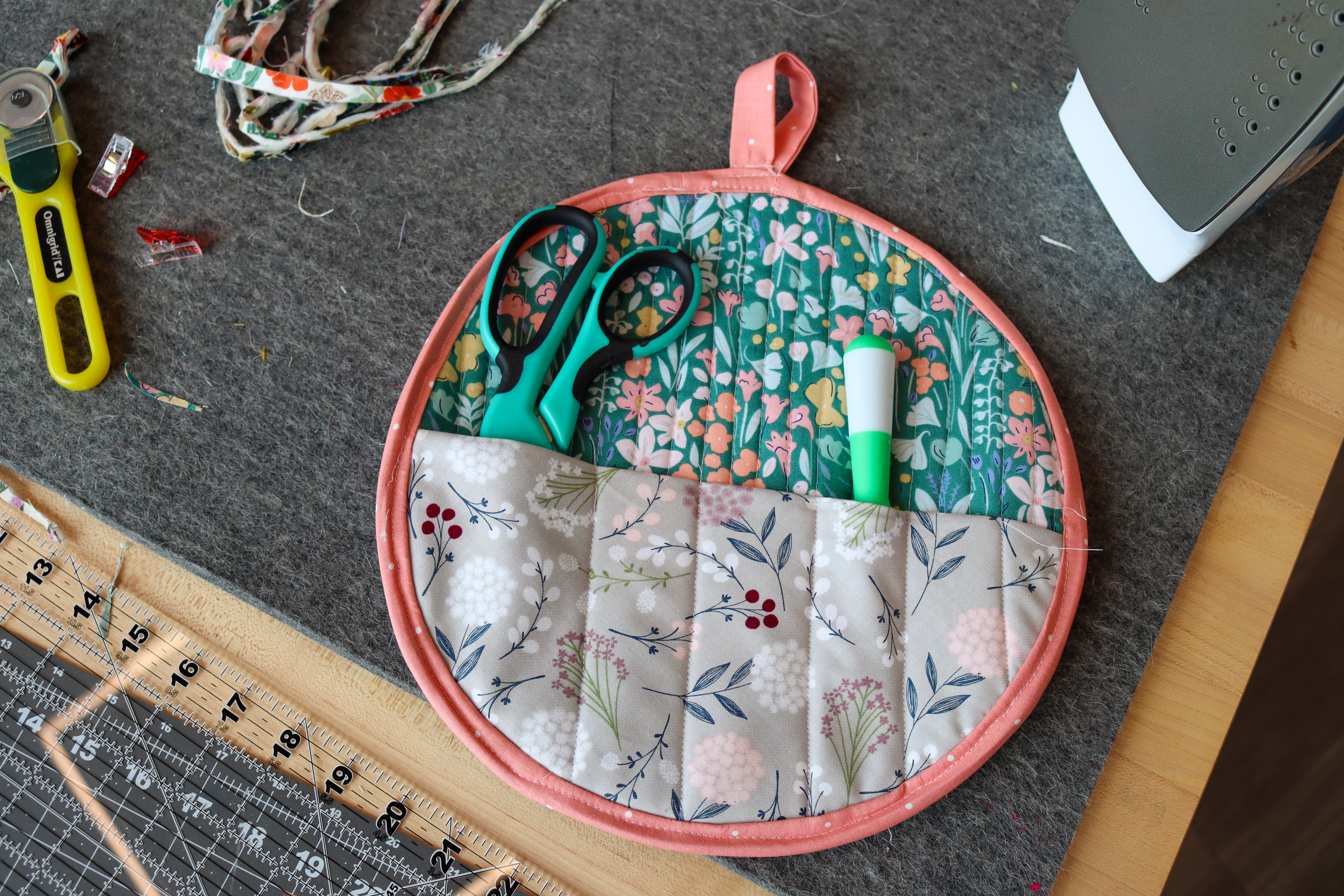Circle made of quilted floral fabric with pouches holding scissors and a marker. There is a loop at the top to hang the organizer.