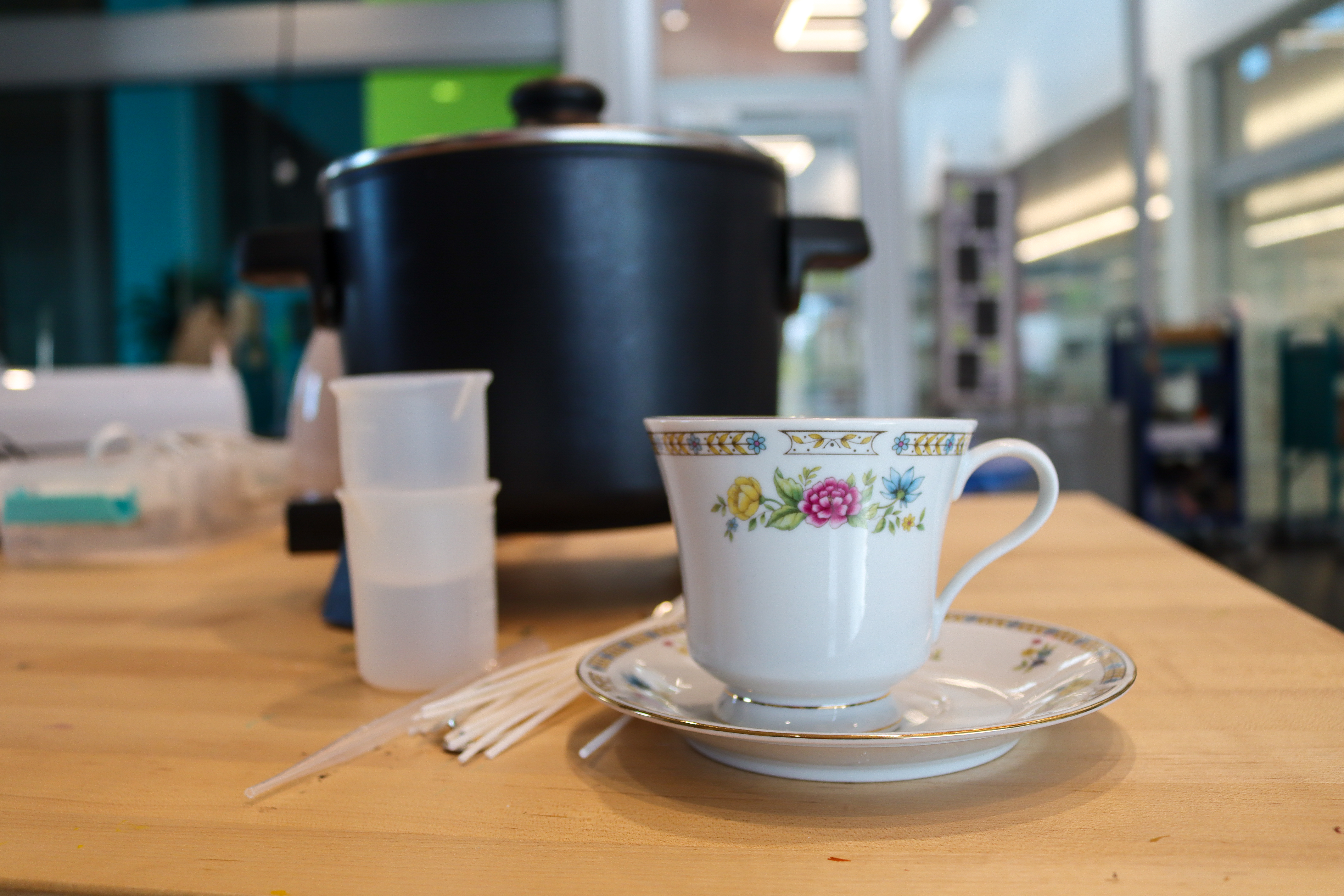 White teacup and saucer with pink flowers on a table next to candle making supplies including a pile of wicks, a pipette, beakers, and a wax warmer.