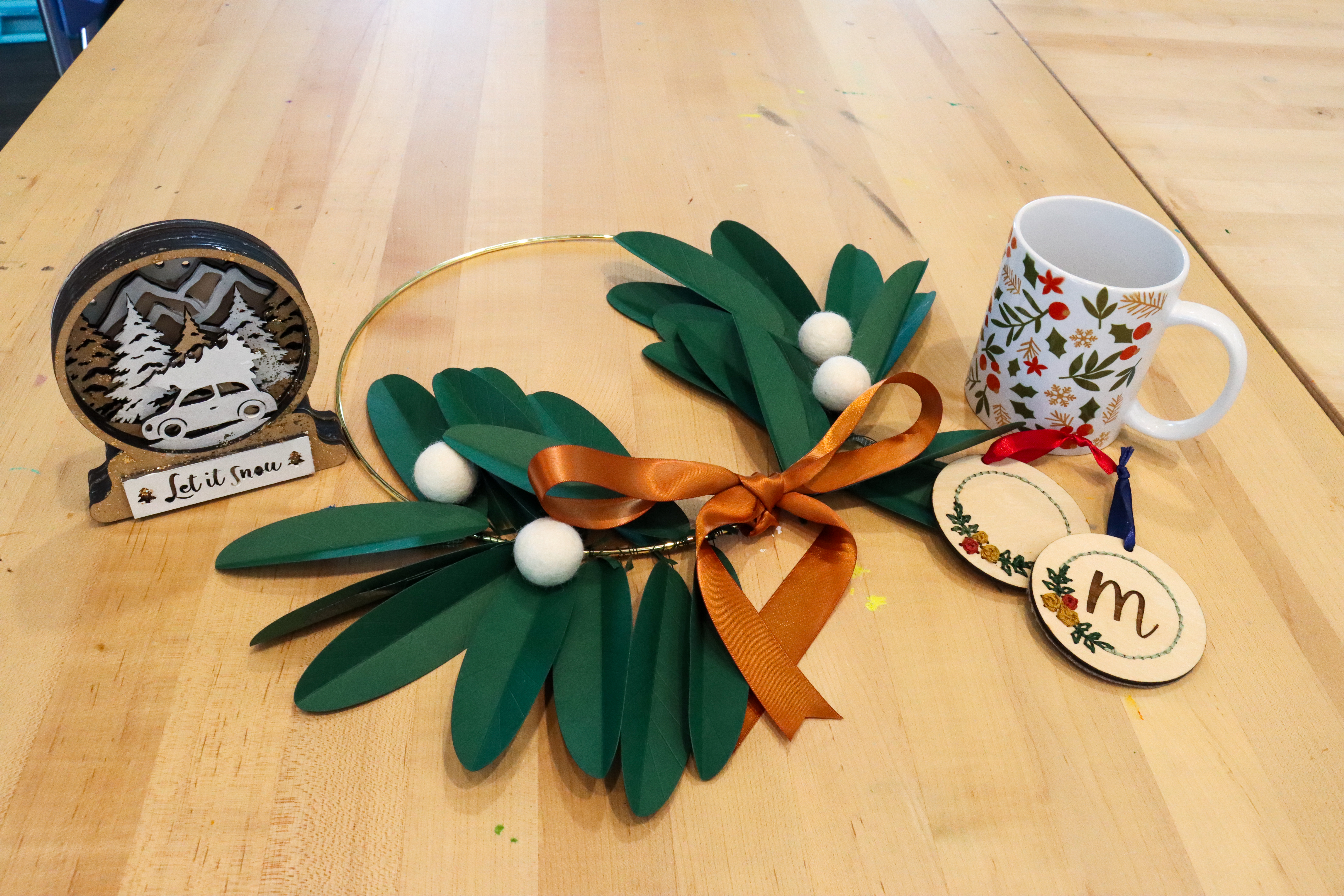 Holiday-themed crafts on a table including a wire and paper wreath, a wooden snow globe, a holiday printed mug, and wooden circular ornaments. 