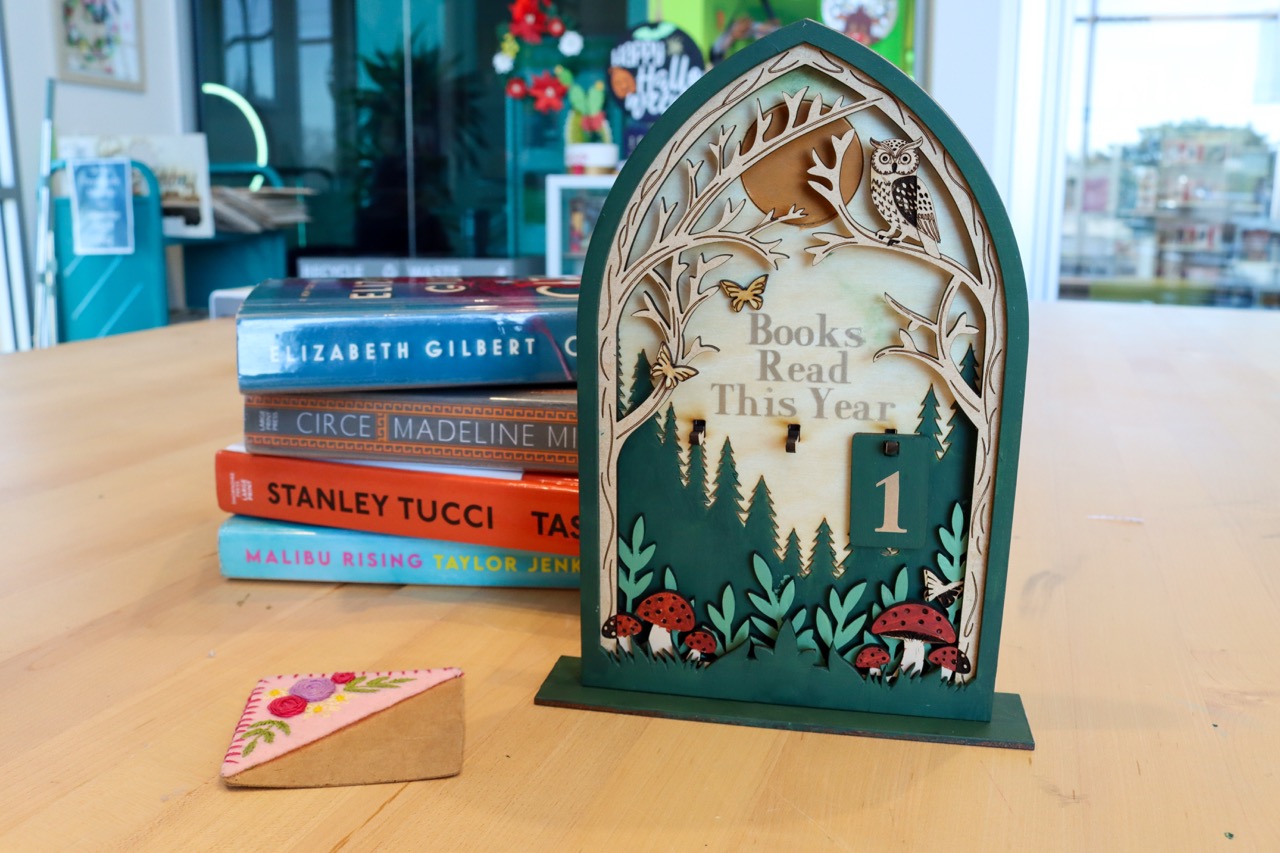 Wooden stand with a forest scene that says books read this year with a counting tracker on a peg. Next to it is a corner bookmark with embroidered flowers on it.