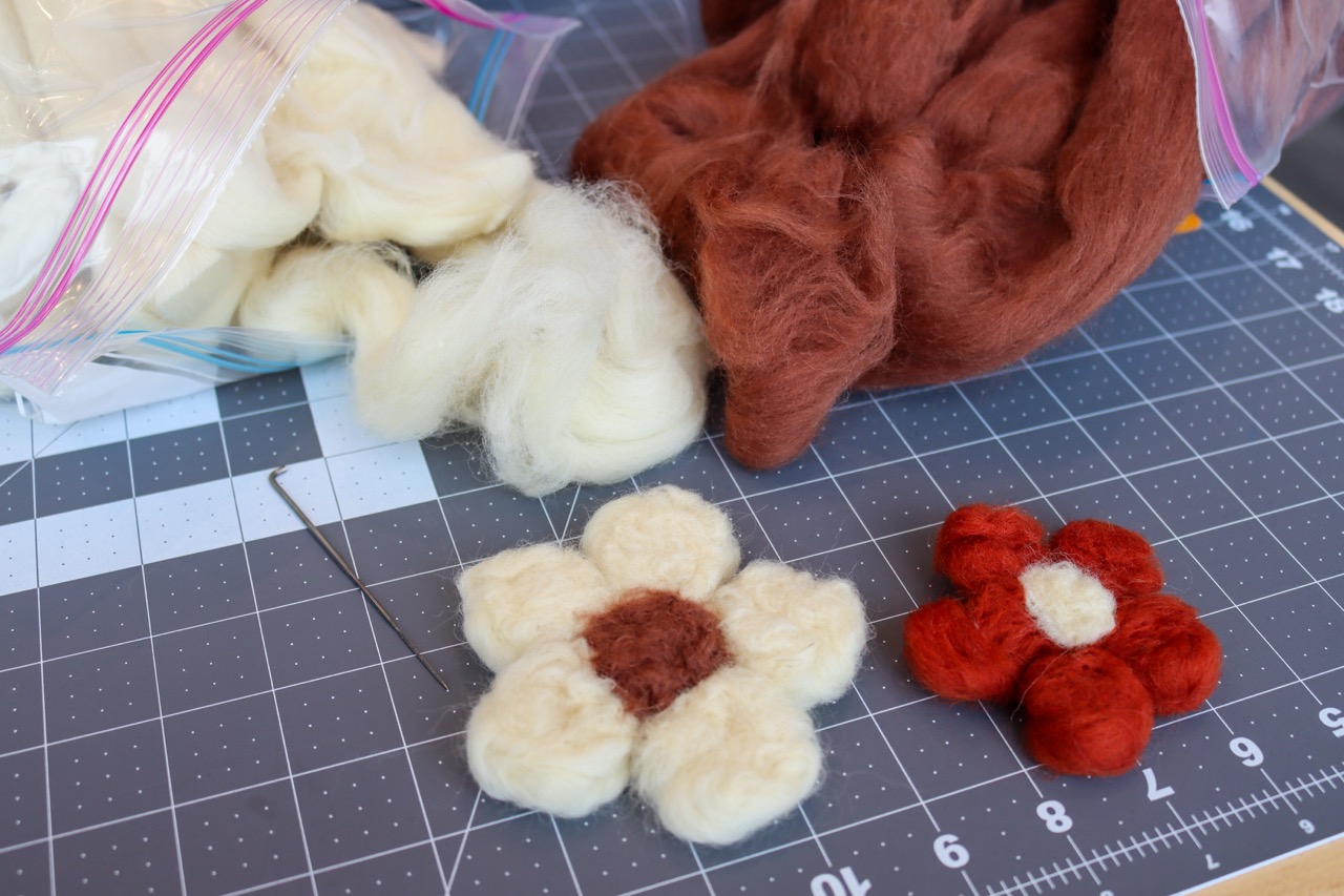 Two felted flowers on a table next to bags of felt and a felting needle.