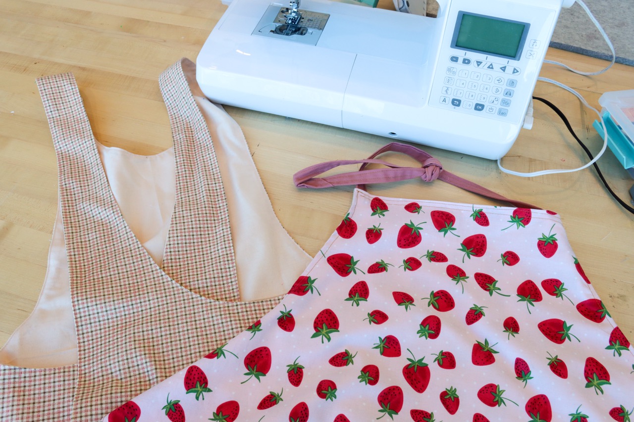 Two aprons laying on a table, one with a traditional tie neck, and the other a cross-back.