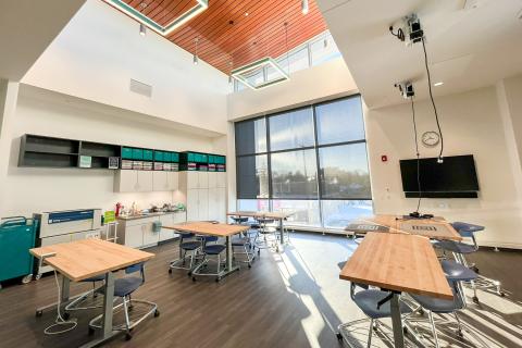 Studio space with large windows and natural light, gray floors, wooden tables and lofted ceiling with wood panelling.